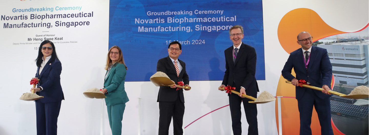 Five people in suits holding shovels with dirt at a Novartis groundbreaking ceremony.